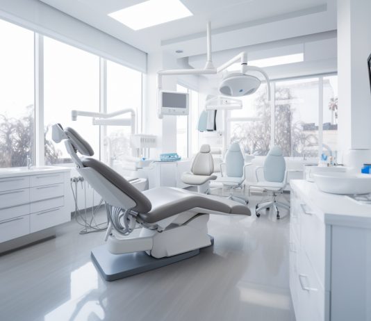 Modernes und helles Behandlungszimmer in einer Zahnarztpraxis mit dentaler Ausstattung, darunter ein Zahnarztstuhl, Leuchten, Monitore und medizinische Geräte. Große Fenster lassen viel Tageslicht herein.