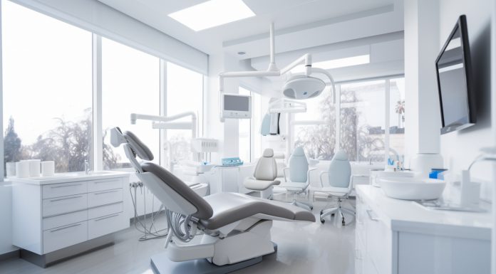 Modernes und helles Behandlungszimmer in einer Zahnarztpraxis mit dentaler Ausstattung, darunter ein Zahnarztstuhl, Leuchten, Monitore und medizinische Geräte. Große Fenster lassen viel Tageslicht herein.